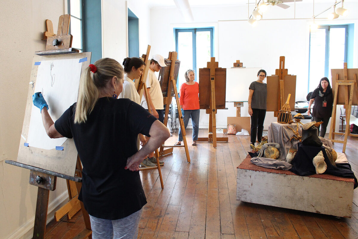 Six students look at an assortment of objects while attempting to blindly draw them on easels with their left hands.