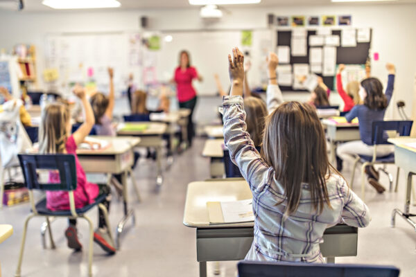 Elementary school students raise their hands while sitting at their desks.