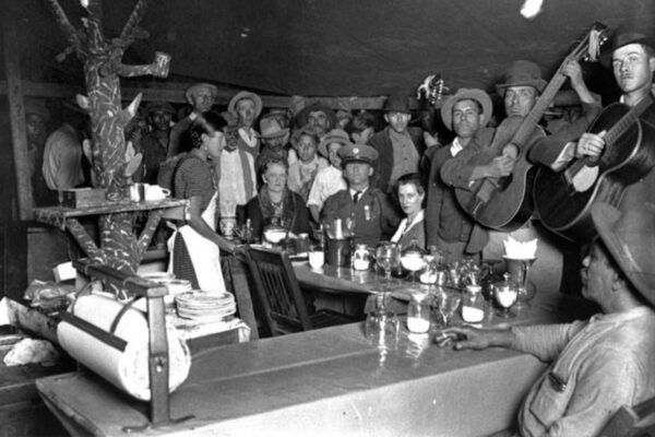 Women, children and musicians stand around a table listening to music.