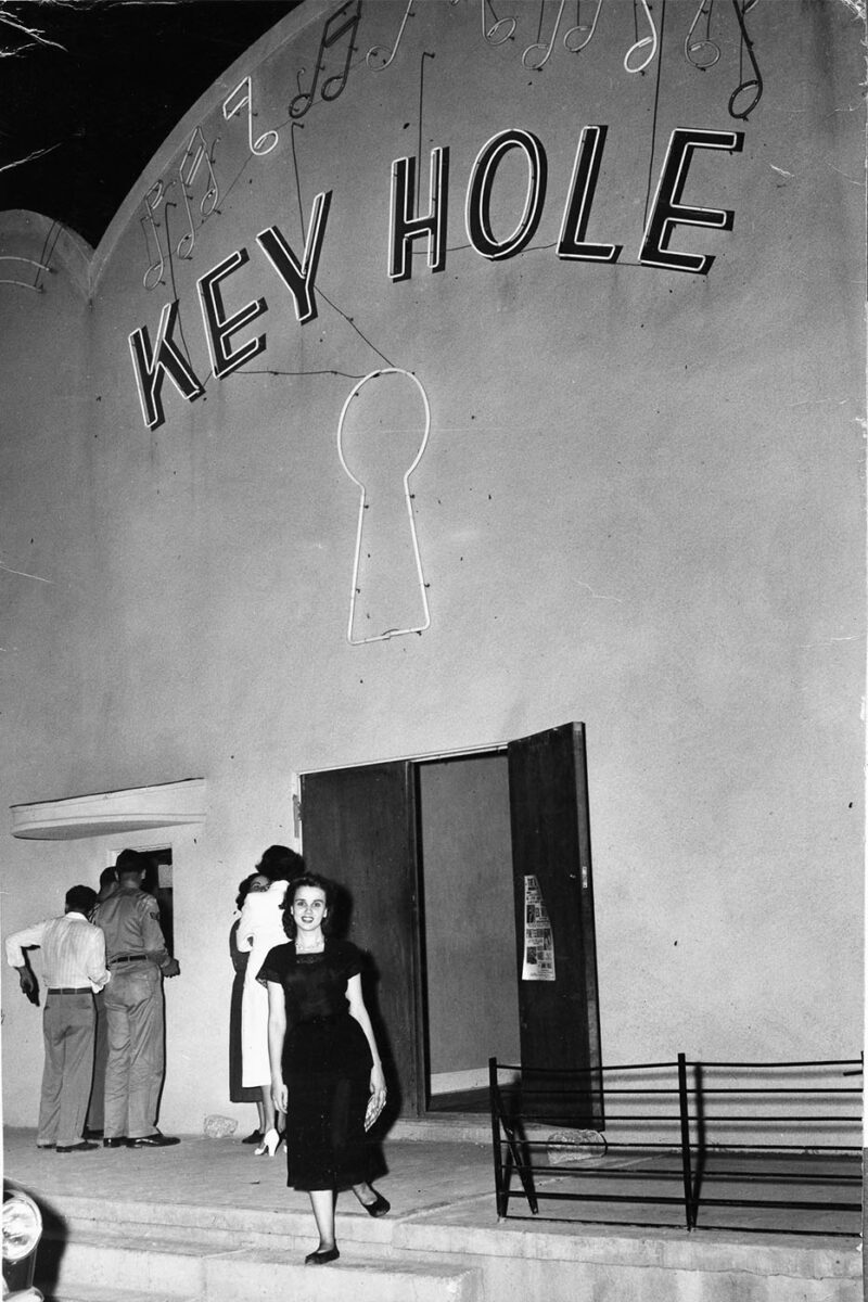 A woman in a dress stands on the front steps of the Keyhole club.