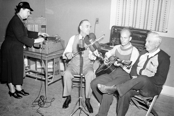 Three men sit in a room playing the guitar and violin as Ruby Lomax records the music,