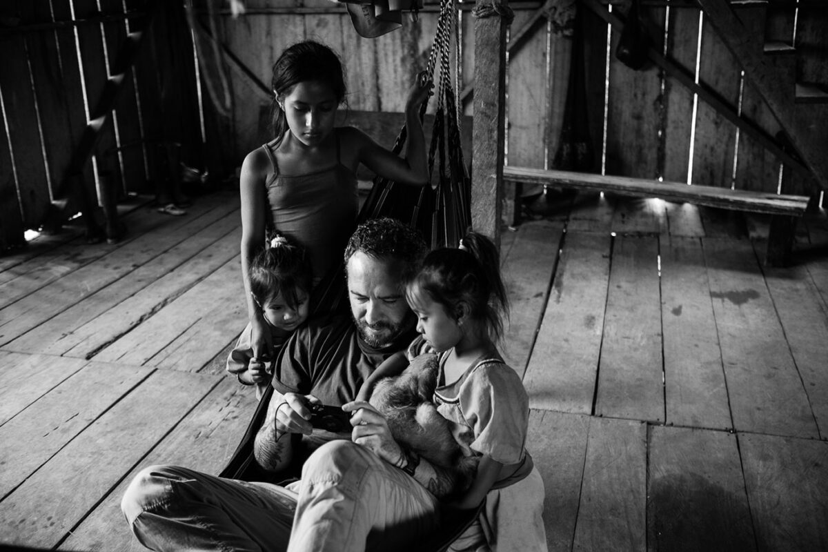 Michael Cepek on a hammock in a hut showing pictures on his phone to three young girls