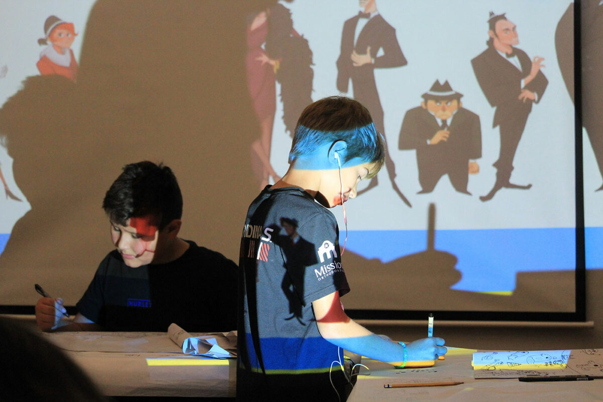 Two young boys stand in front of a projector while they draw