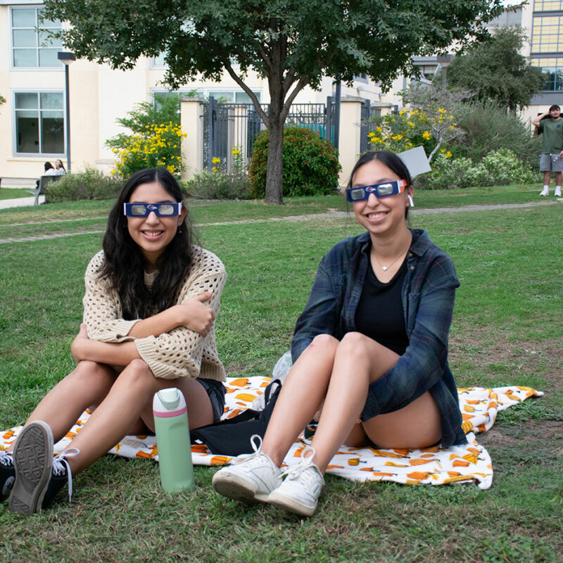 Two students sit on blankets on a lawn wearing solar eclipse glasses.