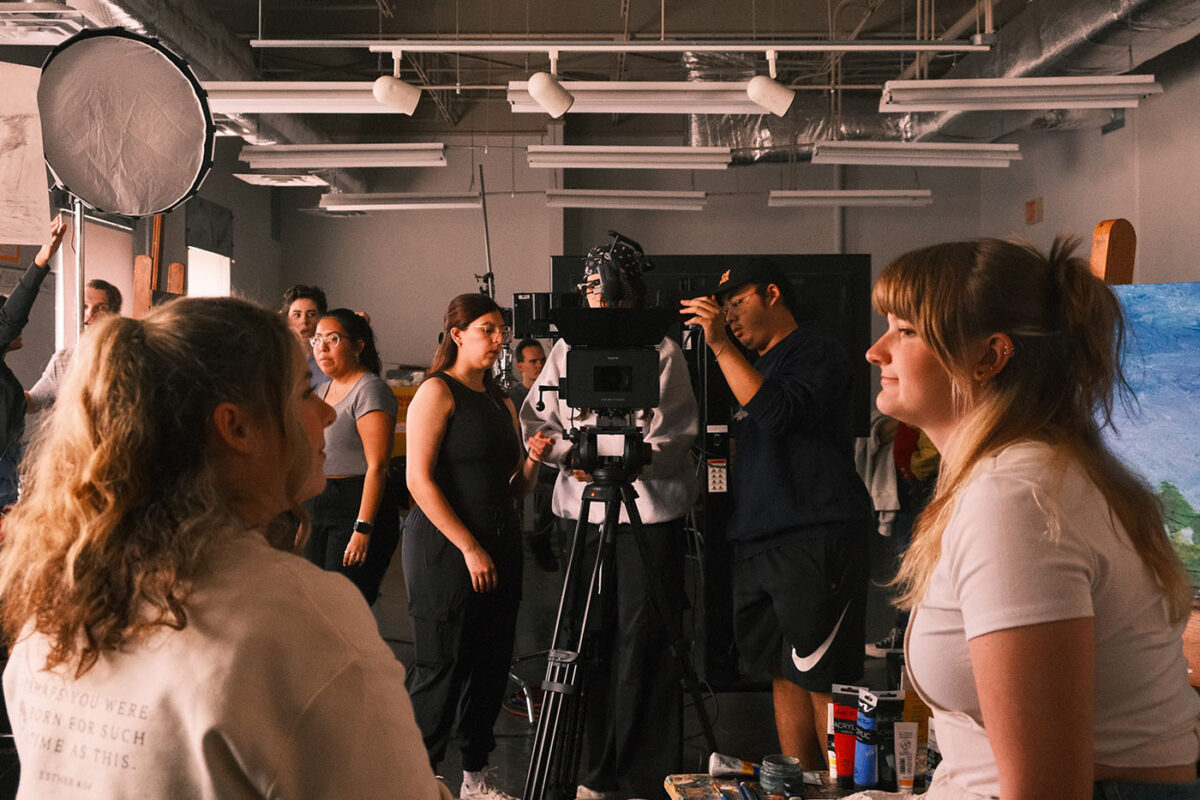 Two actors sit in the foreground while a film crew prepares for the scene in the background