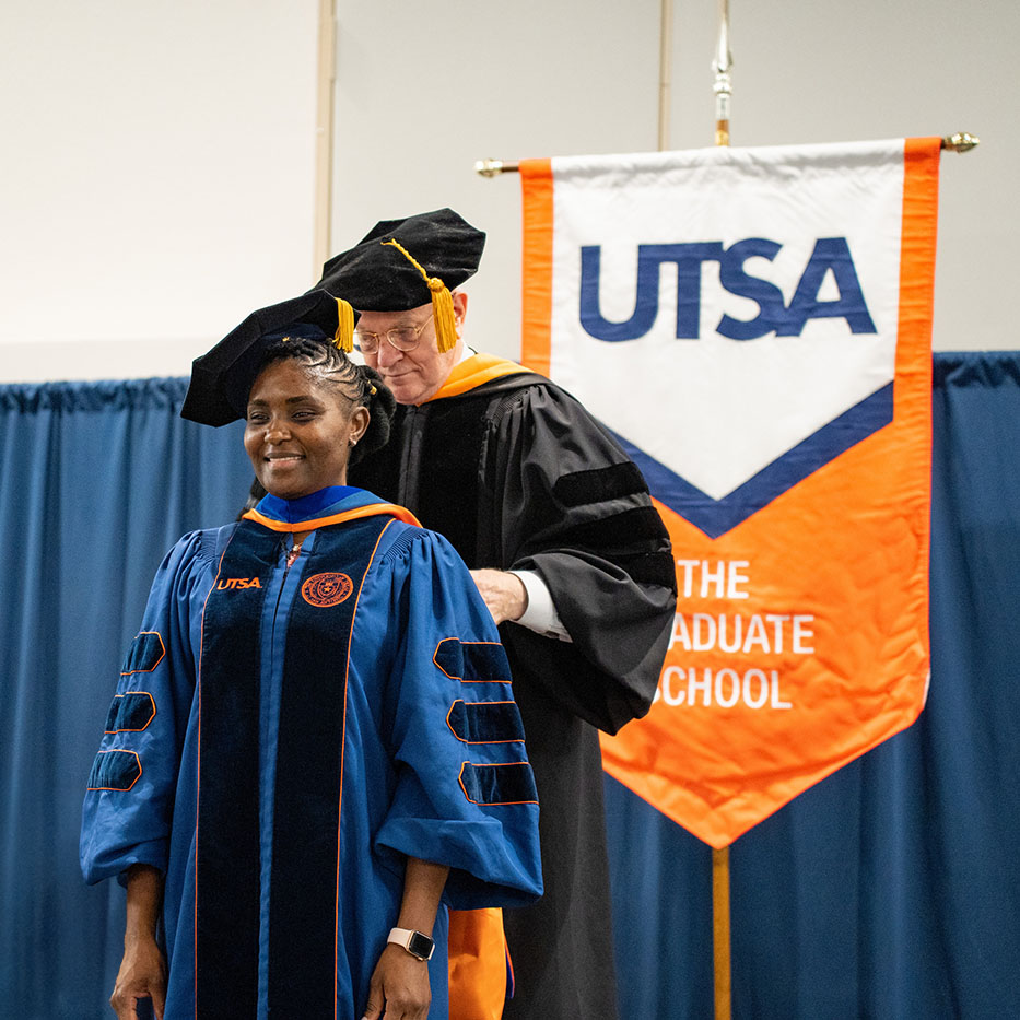 A man places a hood on a UTSA doctoral graduate