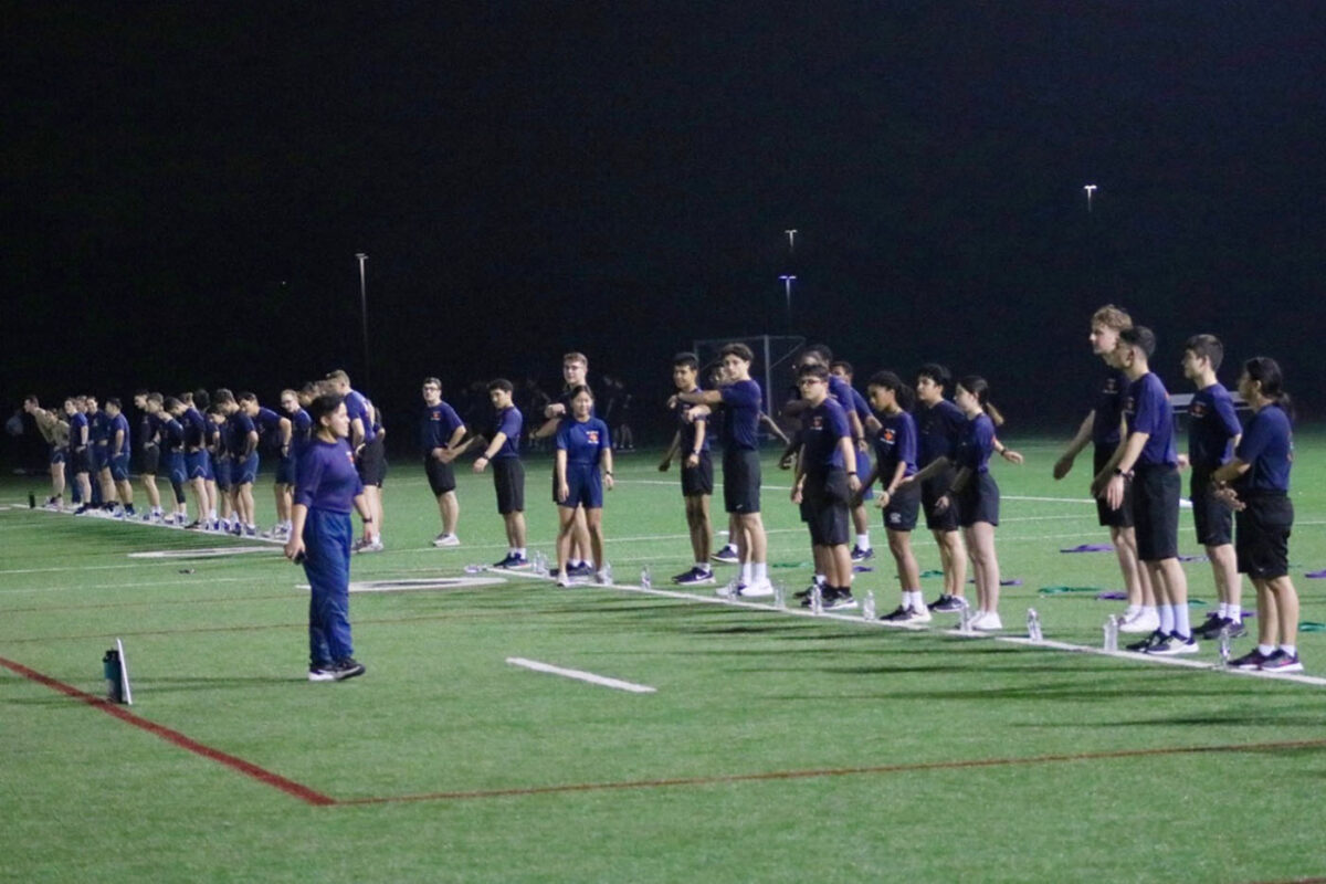 A row of cadets in navy blue t-shirts and black shorts stretch their arms while an instructor watches on.