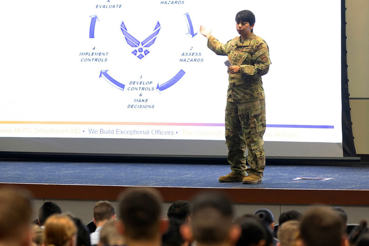 Major Tina Cavazos speaks on stage while pointing to a presentation on a projector screen.