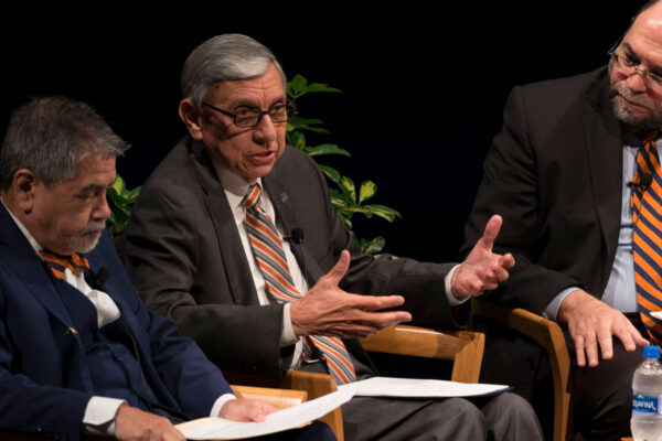 Three men sit side by side having a conversation on a stage.