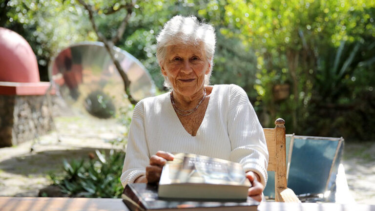 A woman with white, short hair sits with a stack of books and looks straight ahead.