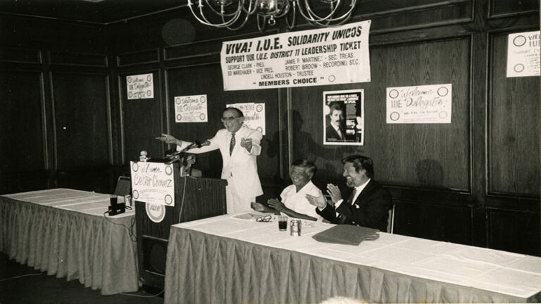 A man in a white shirt stands at a podium with his arms out while two men sit at a table beside him.