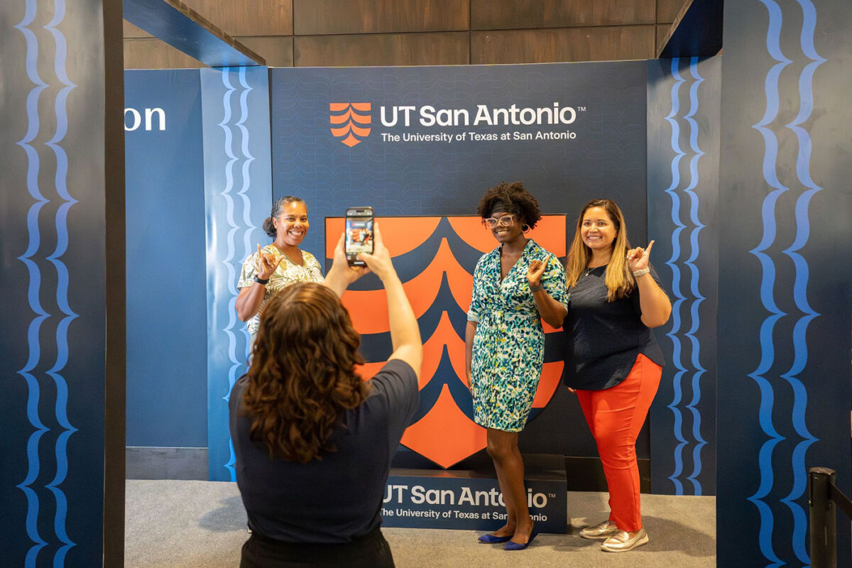 Three women stands beside a large orange shield and as they make a gesture with their hand, which symbolizes a roadrunner.
