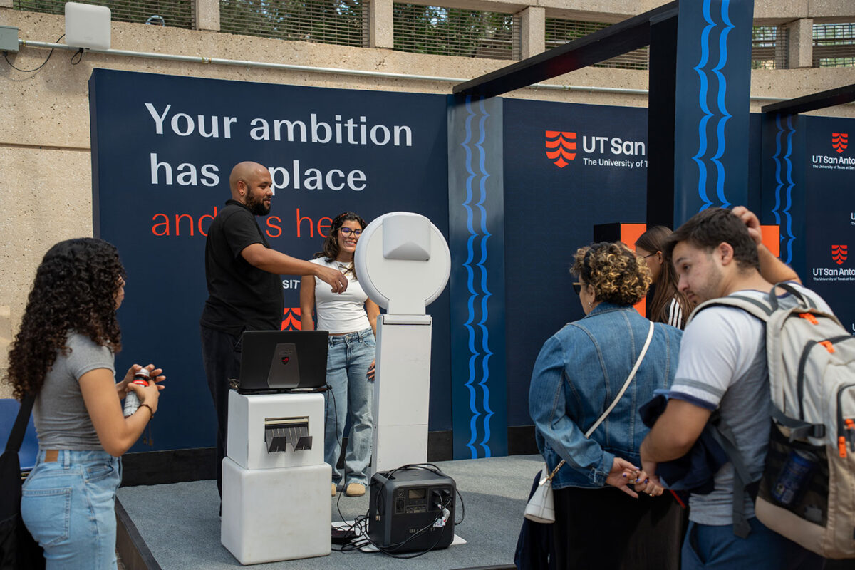 A woman smiles in front of a white camera while a man assists in taking the photo. Other people wait in line to take a photo.