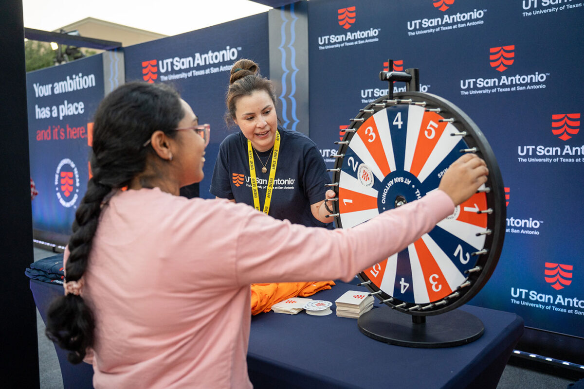 A lady in a blue shirt stands behind a table as another person spins a wheel with numbers.