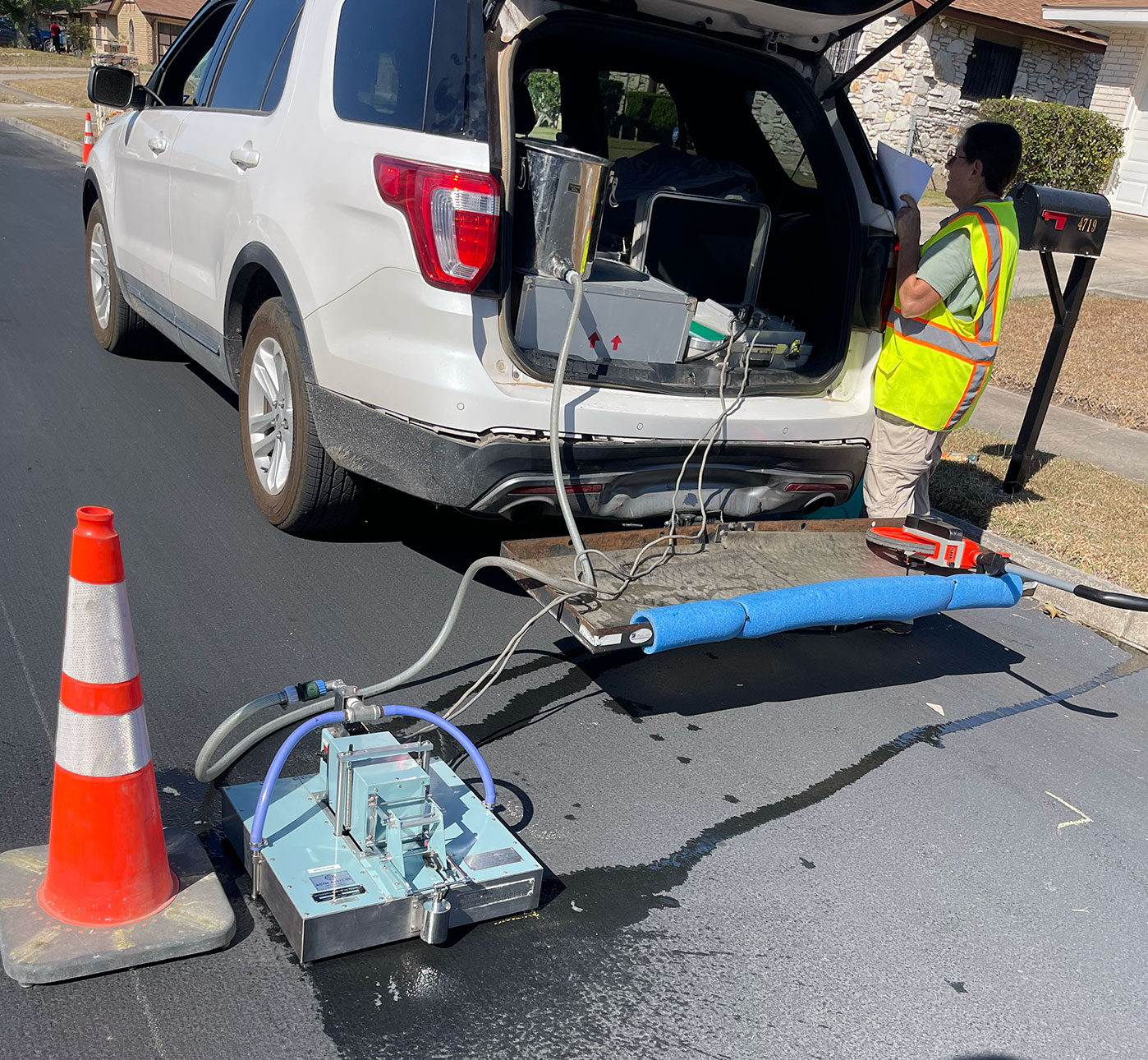 A person in a yellow safety vest stands underneath the open hatch of a SUV with various tools on the road.