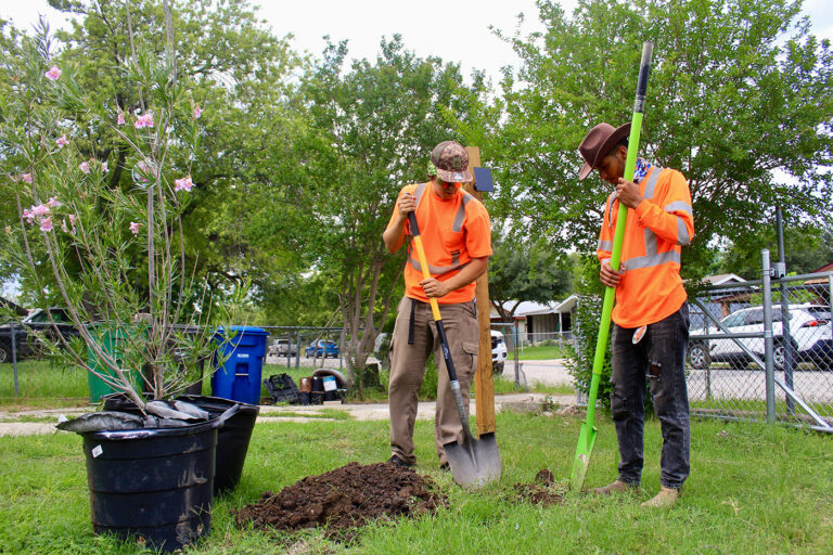 Two men in orange safety shirts use shovels in dig a whole in the ground.