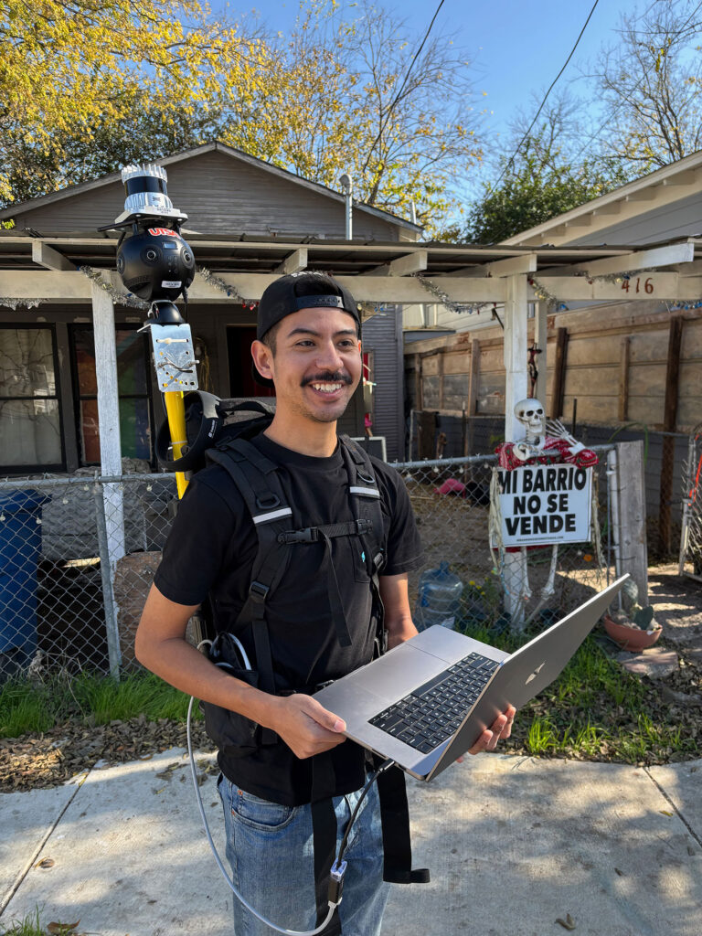 A young man looks at the camera to smile with a laptop in his hand.