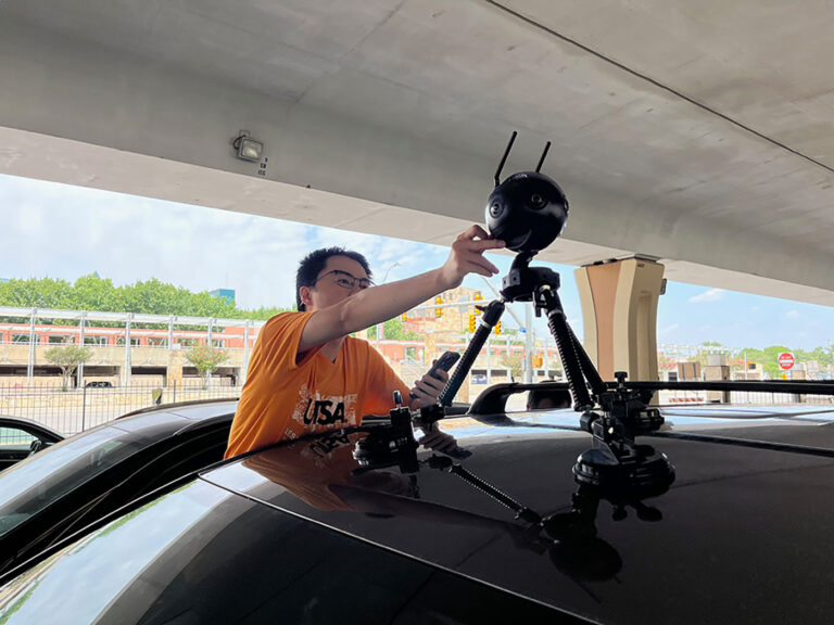 A man stands beside a car to reach up and fix a machine on the roof.