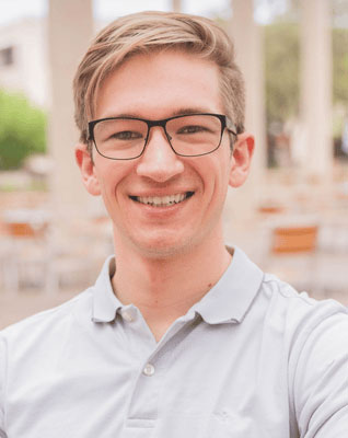 A young man in glasses and a polo stands outside smiling.
