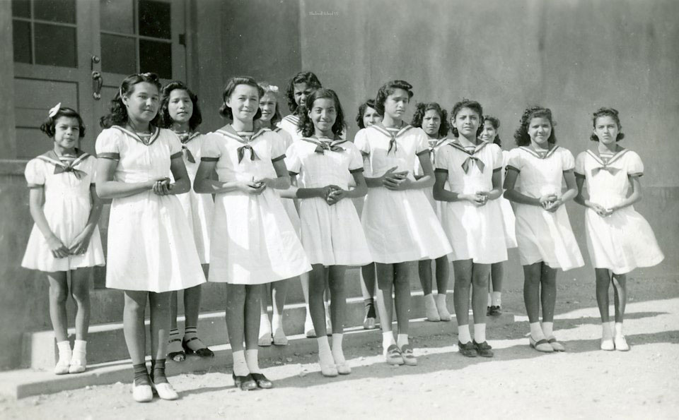 A black and white photo of elementary girls in white dresses with a dark colored collar and bow at the kneck.