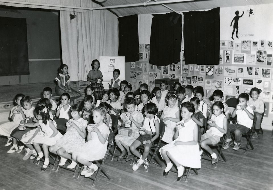 Black and white image of elementary students sitting in a room with a stage. All students are drinking cartons of milk.