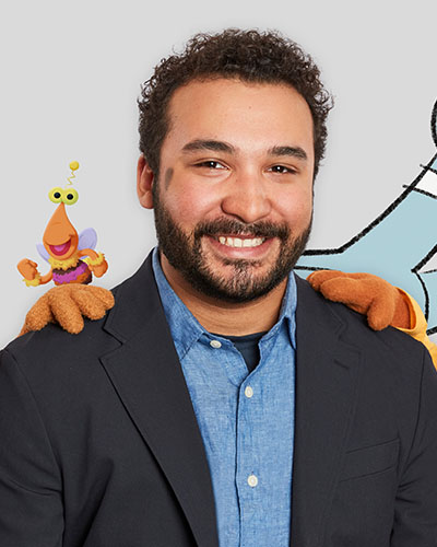 A man in a black jacket and blue shirt smiles at the camera with two puppet hands on each of his shoulders.