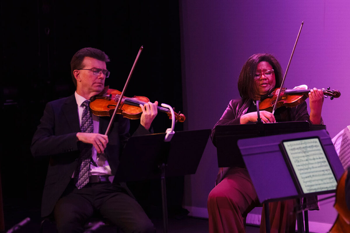 A man and woman sit on stage as they play with violins.