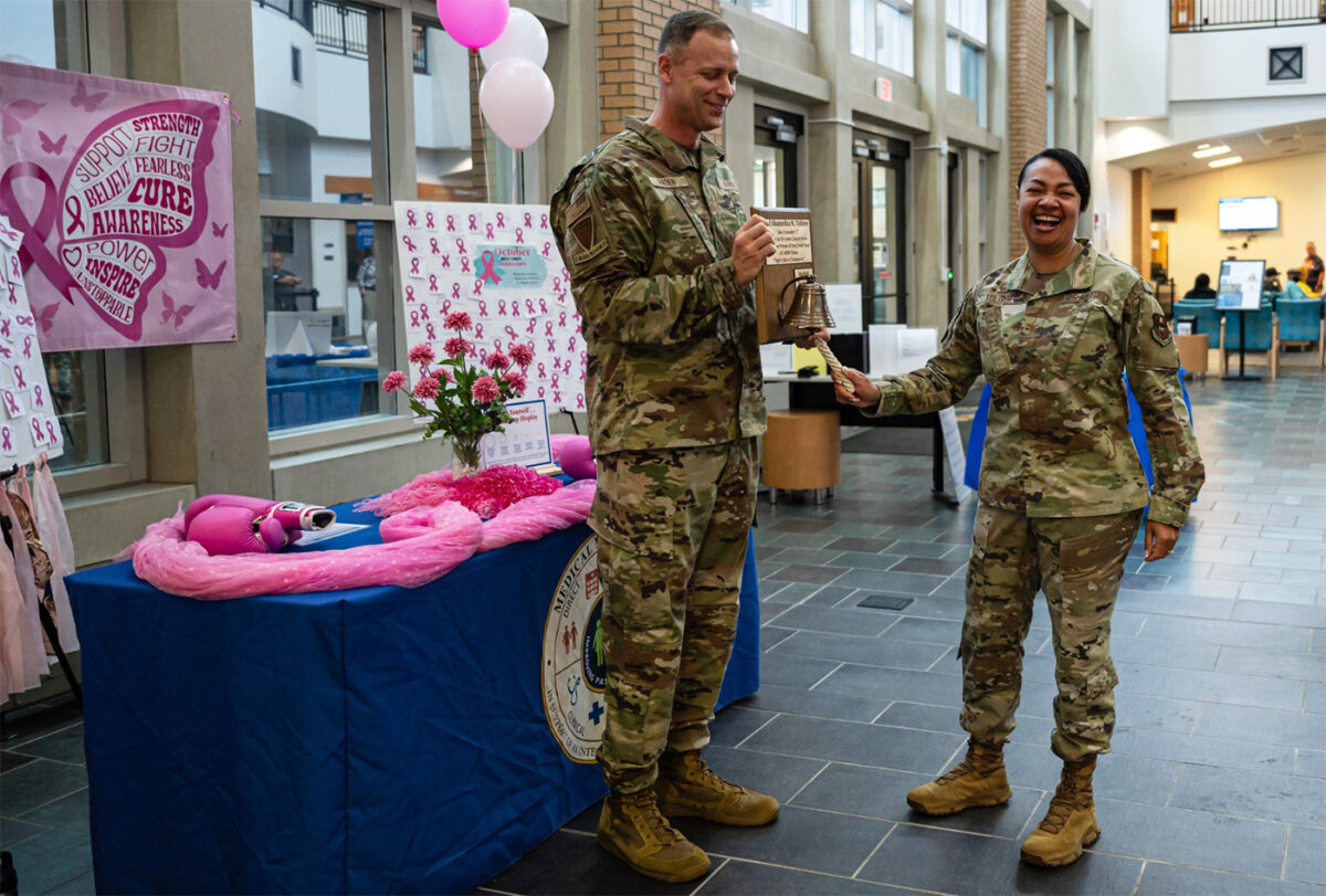 A woman in military gear rings a bell on a plaque a man, also in uniform, is wearing.