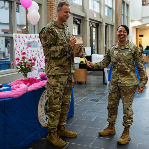 A woman in military gear rings a bell on a plaque a man, also in uniform, is wearing.