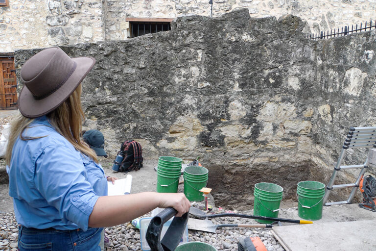 A woman in a tan hat and blue long sleeve shirt rests her hand on a shovel and looks out towards a rock structure.