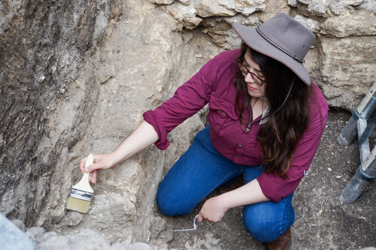 A woman in a hat, jeans and a maroon colored shirt holds a paint brush to dust off a rock structure.