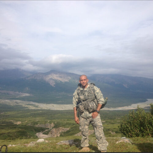 A man stands at the top of a mountain in full military gear.