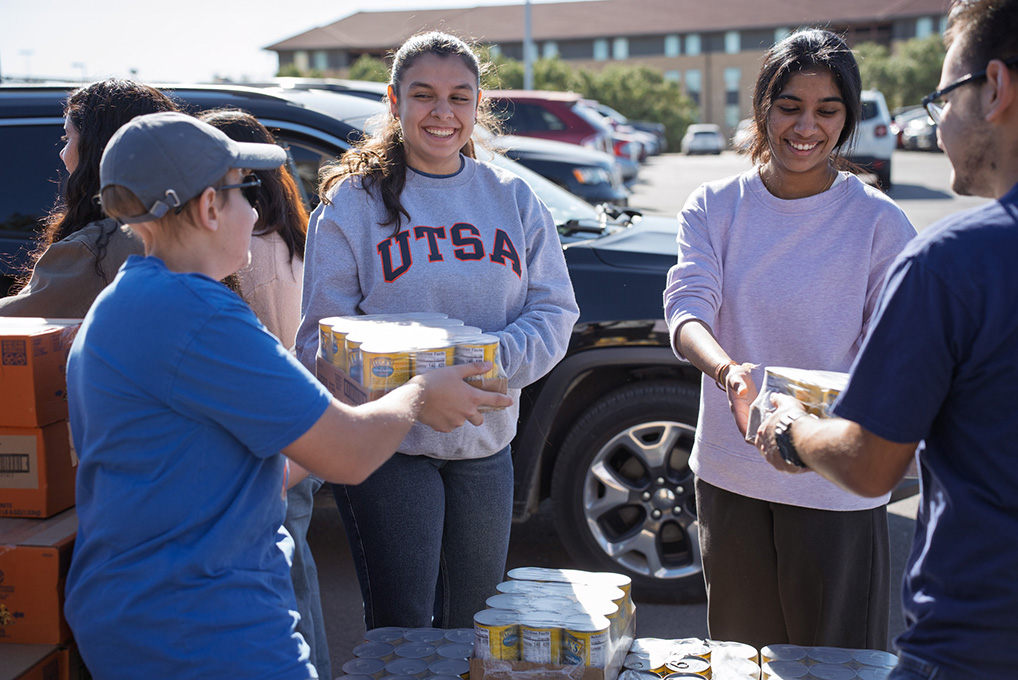 Two women in sweaters hand flats of canned goods to other two people