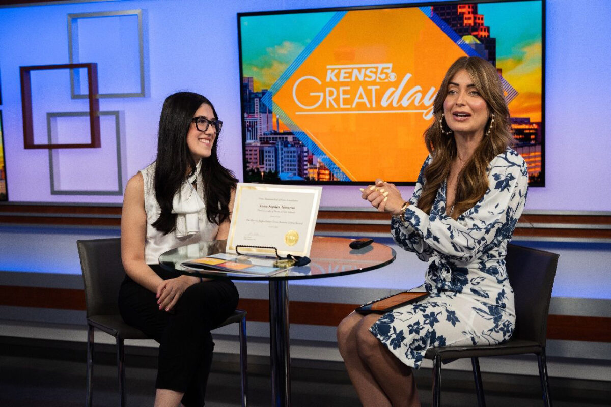 Two women sit at a high-rise table on the set of a tv station as they talk during an interview.