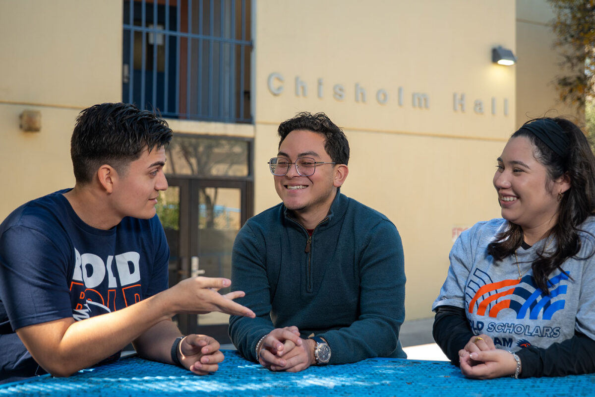 Three people sit at a table table outdoors smiling and talking together.