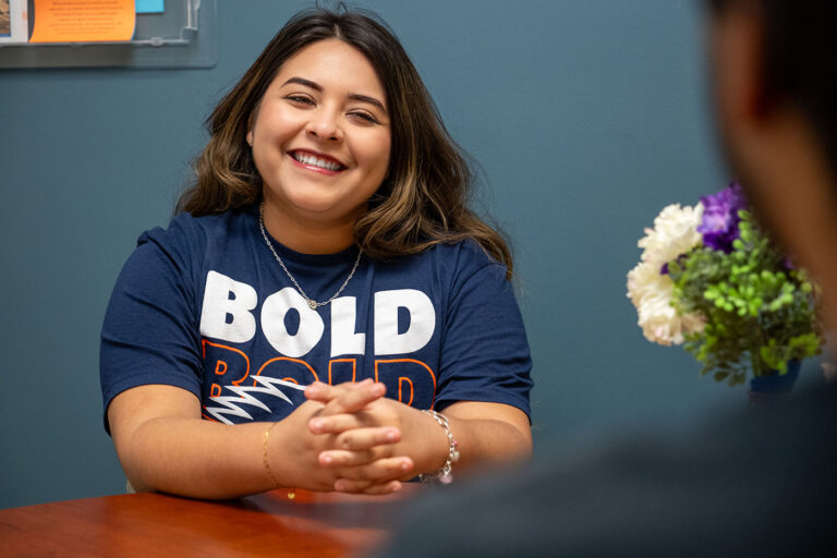 A woman in a blue shirt sits at a table smiling.