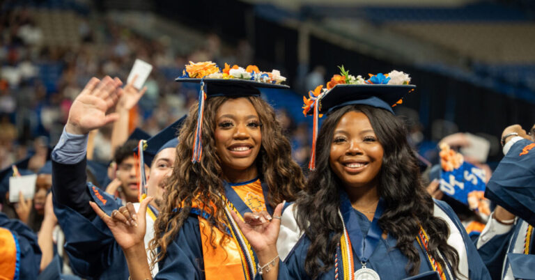 Two students dressed in blue gowns with orange stoles and blue mortar boards on their heads raise their hands up to give a roadrunner hand gesture.