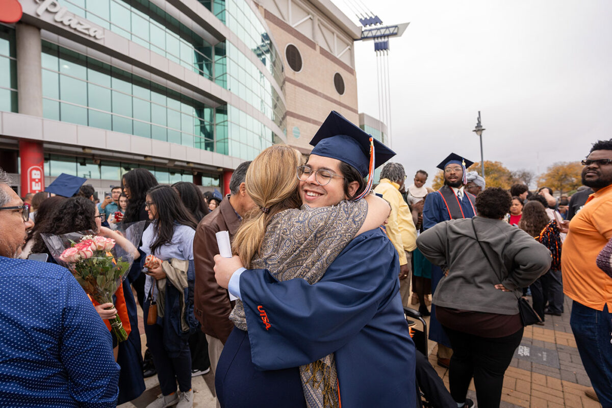 A person in a blue graduation gown and hat hugs a woman outside of a large event center.