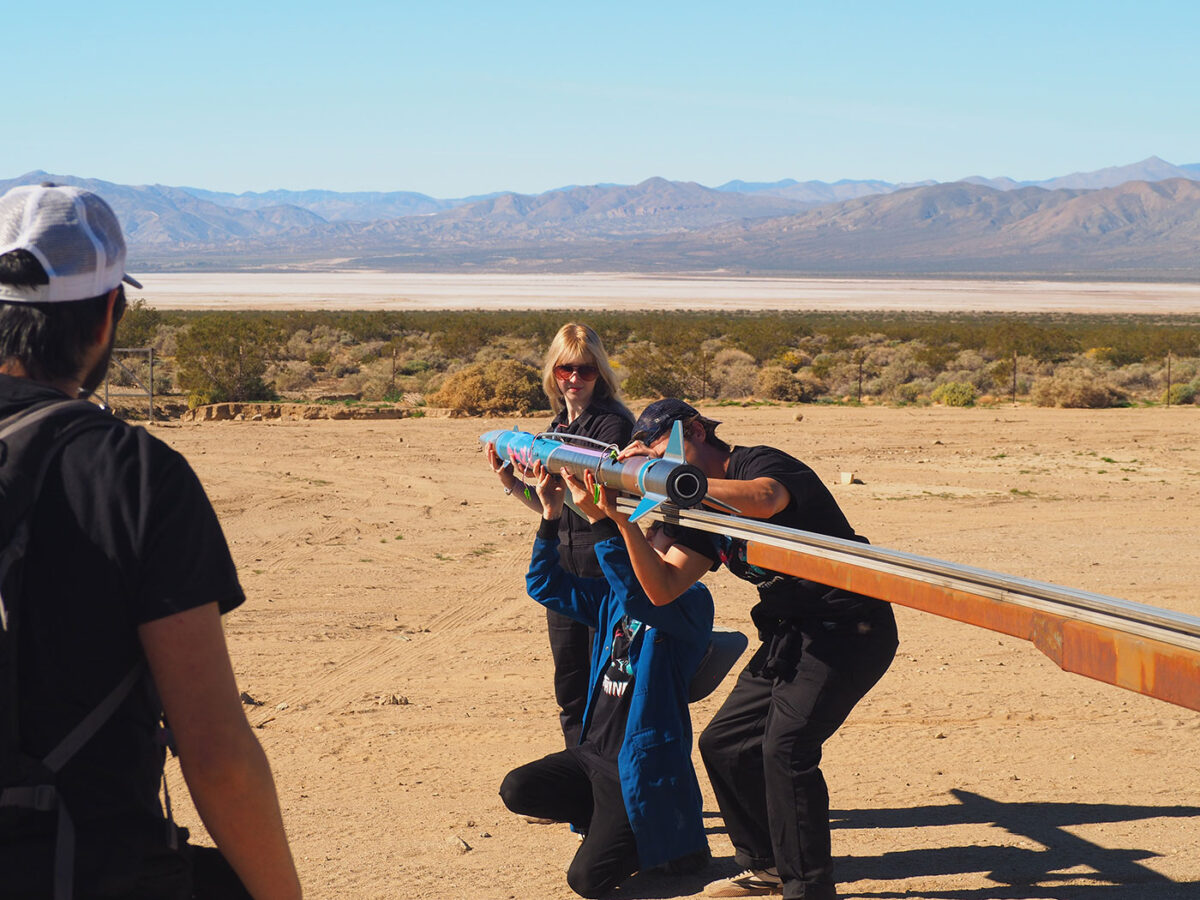 Three people hold a rocket into place on a beam.