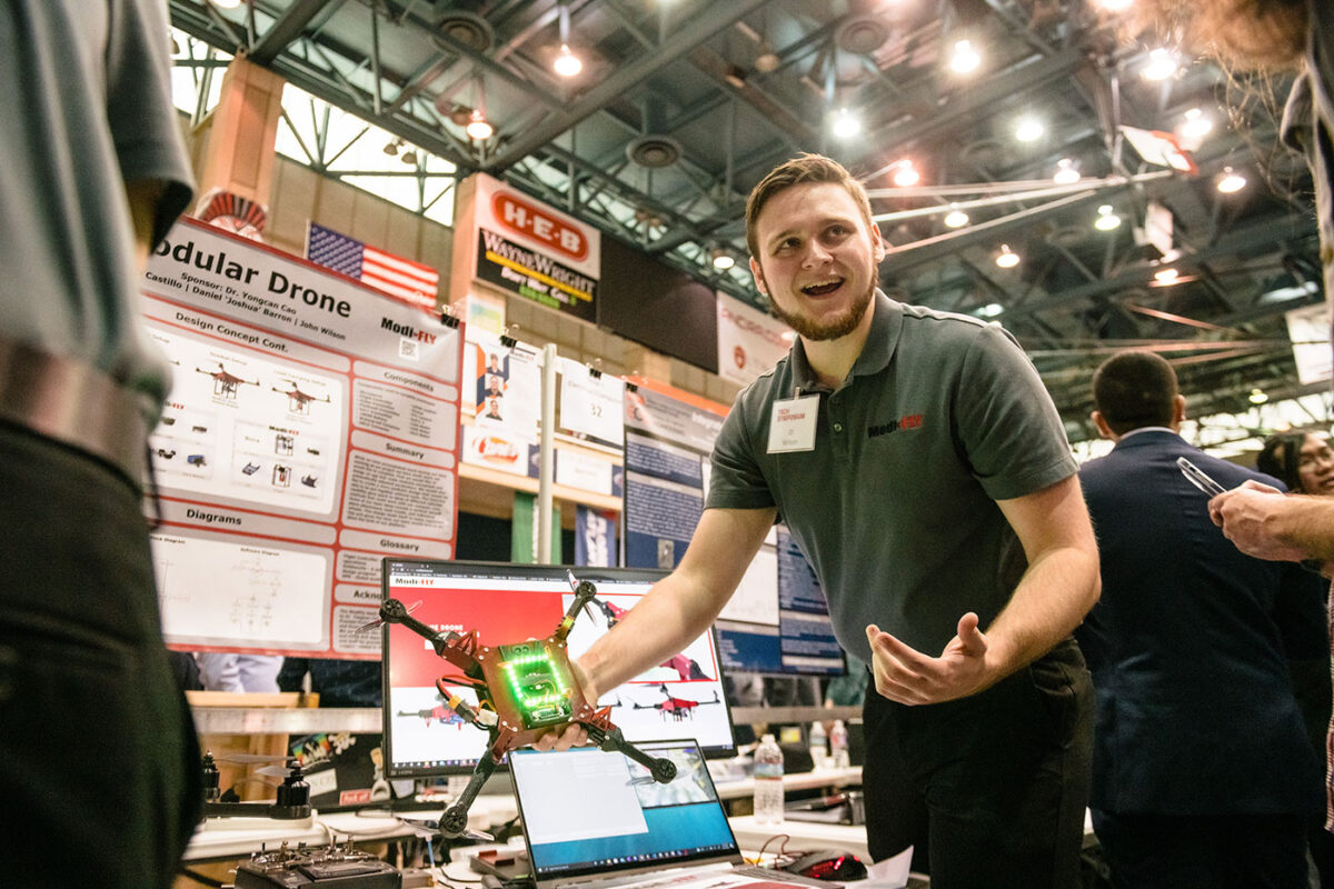A man stands with a piece of technology as he is surrounded by posters of research.