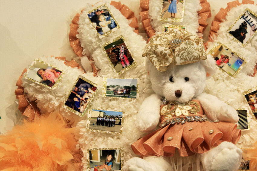 A closeup of a homecoming mum with a large teddy bear, which has an ornate bow and a glittery dress. The bear is surrounded by family photos. This is part of the "Mumentous: The Upsizing of a Texas Tradition" exhibit at the Institute of Texan Cultures.
