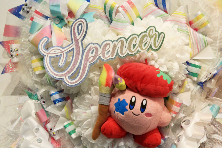 A closeup image of a homecoming mum with the name "Spencer," surrounded by white flowers, rainbow ribbons and a plush of the video game character, "Kirby. " This is part of the "Mumentous: The Upsizing of a Texas Tradition."