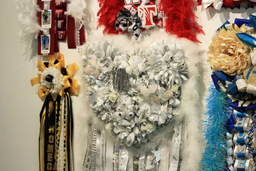 A closeup image of a large heart homecoming mum with white and silver flowres, feathers, bows, ribbons and a teddy bear, which is part of the "Mumentous: The Upsizing of a Texas Tradition" exhibit.