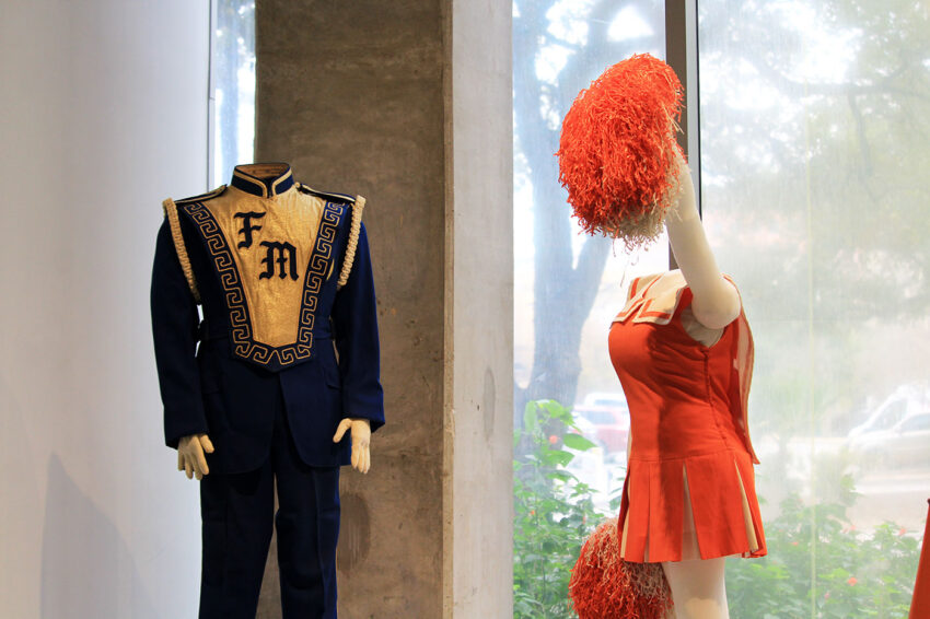 To the left is a a mannequin with a blue high school band uniform with white embroidered details and to the right is a mannequin in an orange and white cheerleader uniform, which is part of the "Mumentous: The Upsizing of a Texas Tradition" exhibit at the Institute of Texan Cultures.