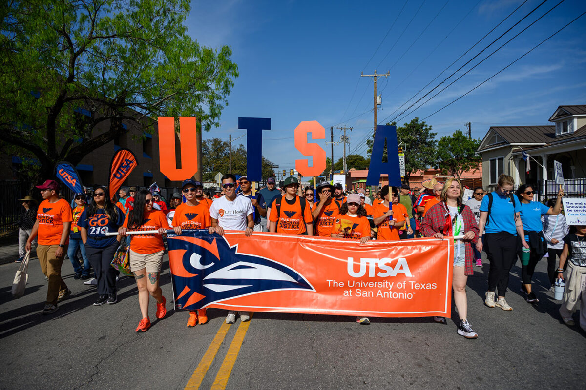 People hold up a large orange banner with a roadrunner image as they walk down a street.
