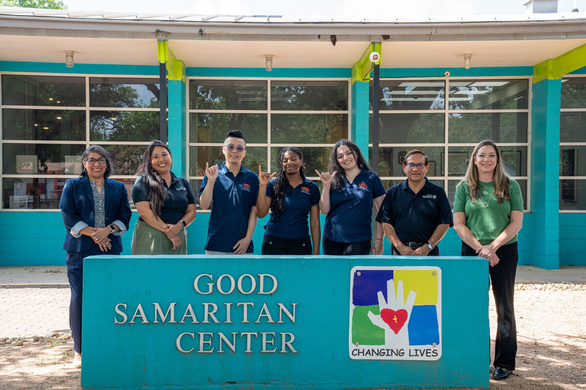 Several people in blue shirts stand in front a sign that says, "Good Samaritan."