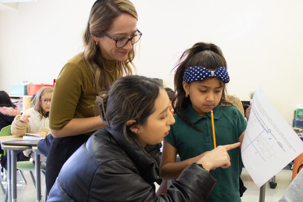 Astrid Venegas helps a student with her classwork while Alejandra Guajardo watches behind them.