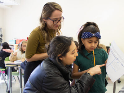 Astrid Venegas helps a student with her classwork while Alejandra Guajardo watches behind them.