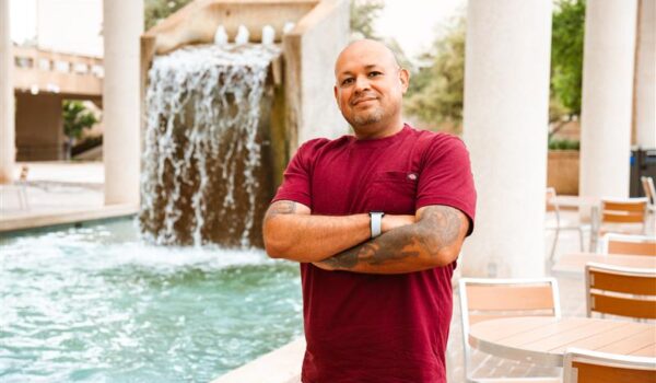 A man in a red shirt and tattoos crosses his arms as he stands in front of a fountain.