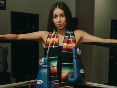 A woman leans against the ropes of a boxing ring. She has boxing gloves hanging from her neck as well as colorful graduation stoles.